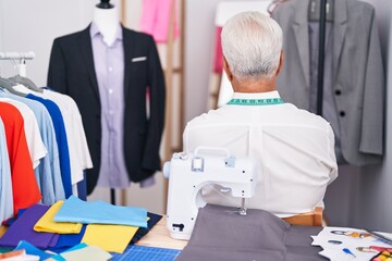 Middle age man with grey hair dressmaker using sewing machine standing backwards looking away with crossed arms