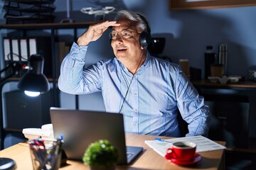 Hispanic senior man wearing call center agent headset at night very happy and smiling looking far away with hand over head. searching concept.