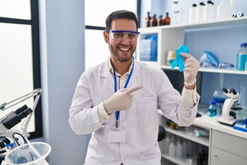 Young hispanic man with beard working at scientist laboratory holding blue ribbon smiling happy pointing with hand and finger