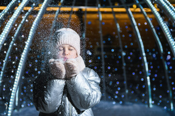 Girl and snow. Girl blowing on palms with snow at night with blue backlight and blurry lights. Holidays theme