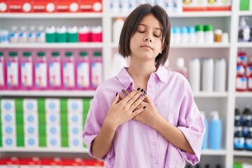 Young girl at pharmacy drugstore smiling with hands on chest with closed eyes and grateful gesture...