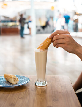 Mano mojando un farton en una horchata en un restaurante de valencia, con fondo desenfocado y en vertical