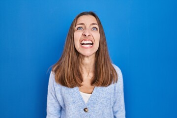 Young woman standing over blue background angry and mad screaming frustrated and furious, shouting...