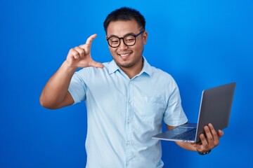 Chinese young man using computer laptop smiling and confident gesturing with hand doing small size sign with fingers looking and the camera. measure concept.
