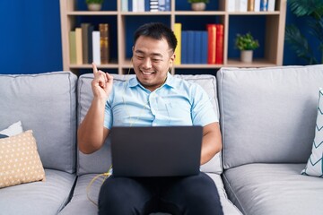 Chinese young man using computer laptop sitting on the sofa smiling happy pointing with hand and finger to the side
