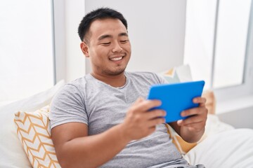 Young chinese man watching video on touchpad sitting on bed at bedroom