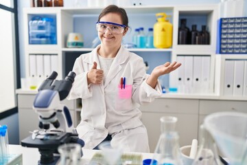 Hispanic girl with down syndrome working at scientist laboratory showing palm hand and doing ok gesture with thumbs up, smiling happy and cheerful