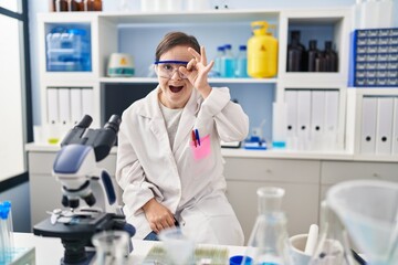 Hispanic girl with down syndrome working at scientist laboratory doing ok gesture shocked with surprised face, eye looking through fingers. unbelieving expression.