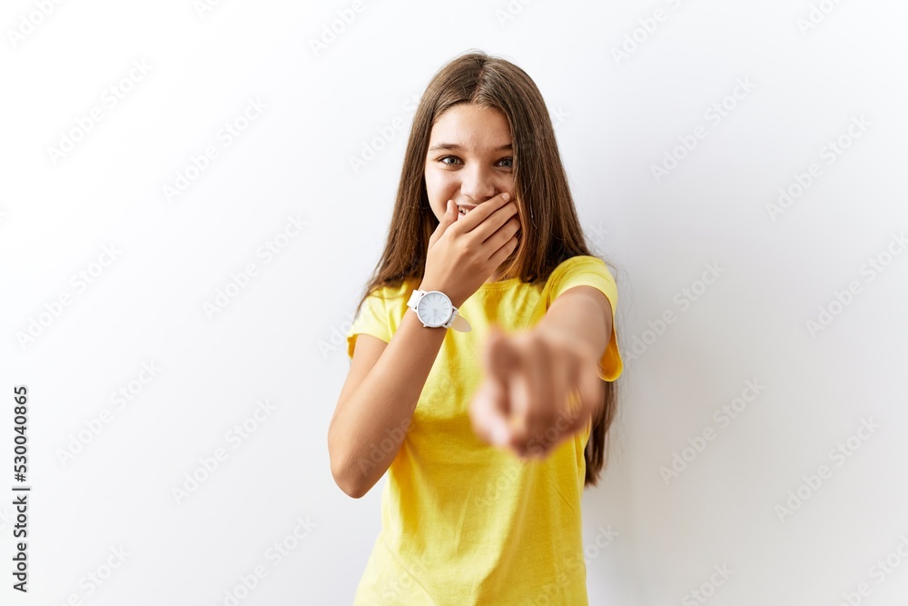 Wall mural Young brunette teenager standing together over isolated background laughing at you, pointing finger to the camera with hand over mouth, shame expression