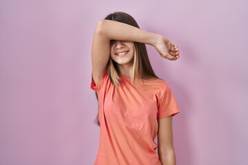 Teenager girl standing over pink background covering eyes with arm smiling cheerful and funny....