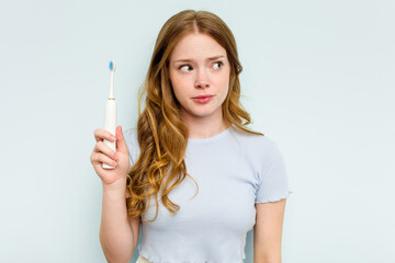 Young caucasian woman holding electric toothbrush isolated on blue background confused, feels doubtful and unsure.