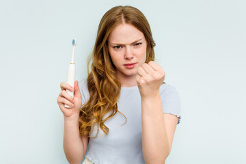 Young caucasian woman holding electric toothbrush isolated on blue background showing fist to camera, aggressive facial expression.