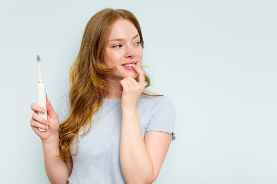 Young caucasian woman holding electric toothbrush isolated on blue background relaxed thinking about something looking at a copy space.