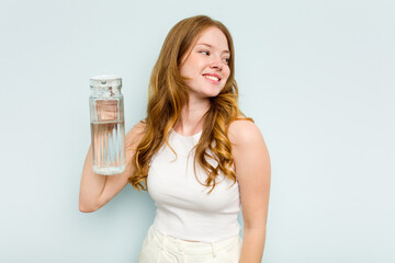 Young caucasian woman holding jar of water isolated on blue background looks aside smiling, cheerful and pleasant.