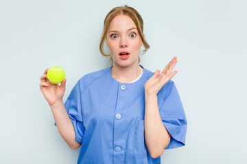 Young caucasian physiotherapist holding a tennis ball isolated on blue background surprised and shocked.