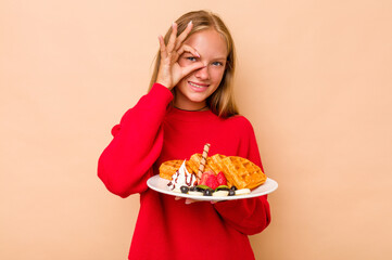 Little caucasian girl holding a waffles isolated on beige background excited keeping ok gesture on eye.