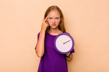 Little caucasian girl holding a clock isolated on beige background showing a disappointment gesture with forefinger.