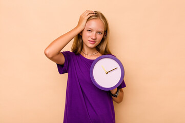 Little caucasian girl holding a clock isolated on beige background being shocked, she has...