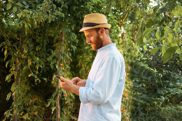 Contemporary agriculture. Bearded Caucasian man in a straw hat using smartphone. The concept of gardening and horticulture