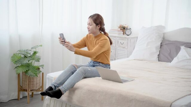 Asian woman sitting on bed taking selfie while using laptop working. Happy female making video call relatives or bestfriend with smartphone at home. Concept of technology.
