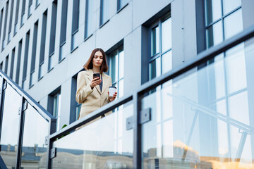 A young brunette girl against the backdrop of a business center, office center. Portrait of a successful startup. Smartphone, coffee casual suit.