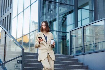 A young brunette girl against the backdrop of a business center, office center. Portrait of a successful startup. Smartphone, coffee casual suit.