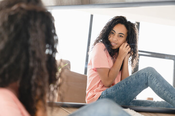 Closeup photo of cheerful young african woman girl putting on earrings, preparing for event meeting date, looking at the mirror at home