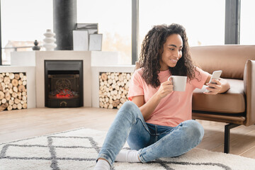 Happy young african woman student freelancer housewife relaxing sitting on the floor reading social media on cellphone, surfing internet, drinking hot beverage at home