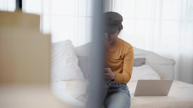 Beautiful asian woman sitting on bed using smartphone chatting with boyfriend with happy, Female wearing hat use free time rest from work on laptop by play iphone in bedroom.
