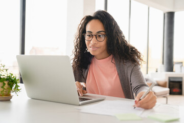 Fototapeta premium Young african student tutor freelancer businesswoman doing homework, paperwork for college, preparing for test exam, using laptop online at home office