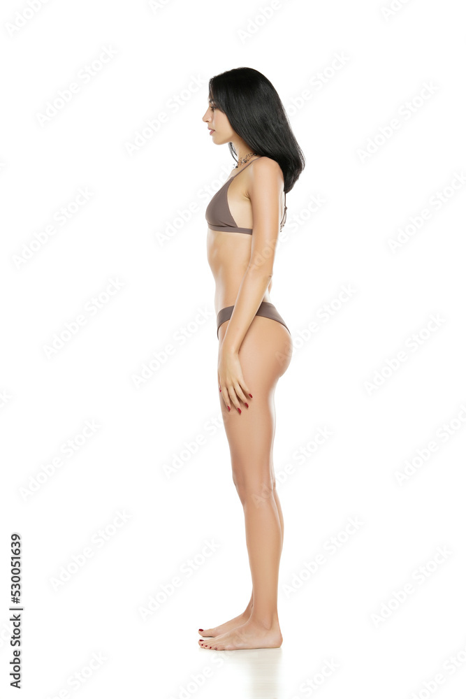 Wall mural side view of a young brunette woman in bikini swimsuit posing on a white background.