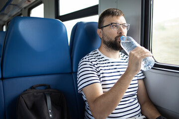 Alone traveler in a train a man drinking water from a transparent plastic water bottle with him next to a backpack in the carriage. Thirst while traveling. Concept: travel, water balance, adventure