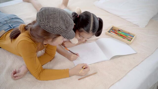 Asian nanny girl spent holiday with cute girl lying on bed painting together in holiday. adorable girl drawing portrait imagination in paper, young girl teaching preschool kid sibling relationship
