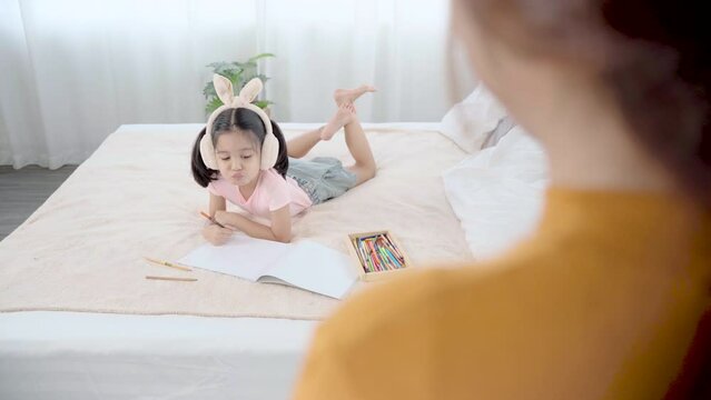 Cute primary school age girls drawing color picture with pencils on bed while mother stand looking with love. Adorable child enjoying creative art hobby activity, children development at home concept.
