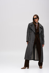 Obraz na płótnie Canvas full length of stylish teenage model in sunglasses and trendy trench coat posing on grey.