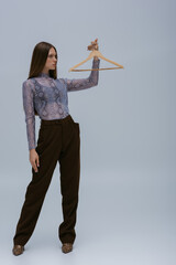 full length of brunette teenage model posing with wooden hanger on grey.