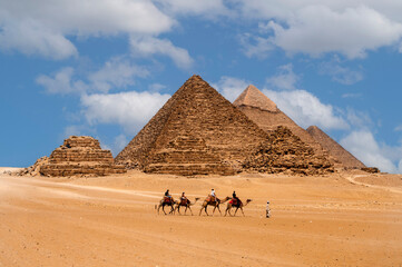Cairo, Egypt. 08.25.2022. Group of tourists riding camels seeing the pyramids of Menkaure, Chephren and Cheops. - obrazy, fototapety, plakaty