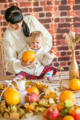 Fototapeta na wymiar A young mother and little daughter in white warm knitted sweaters in an interior decorated with blankets and pillows as well as pumpkins, autumn leaves and apples. Autumn mood. halloween