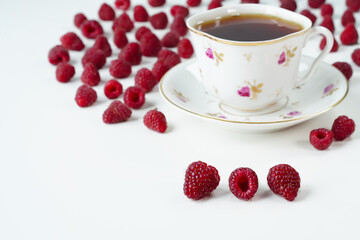 Raspberries on white background, space for text in the bottom left corner, white cup of tea on a saucer with small rose flowers and lots of scattered raspberries in the background, close-up