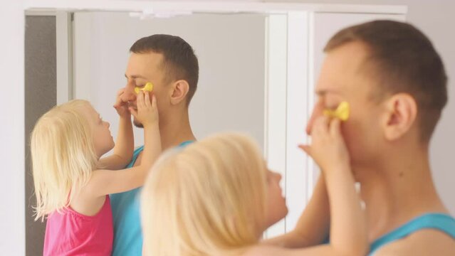 Little daughter puts patches under dad's eyes. Father and blonde child girl having fun in bathroom