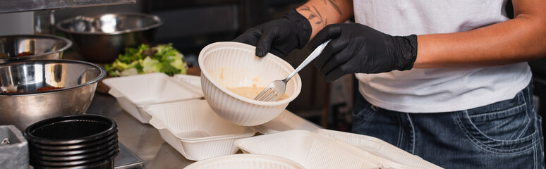 partial view of tattooed african american volunteer holding plastic bowl with soup in kitchen, banner.