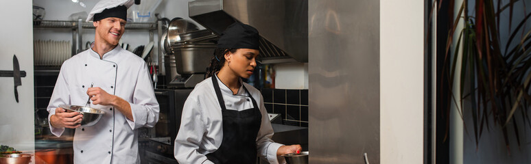 Fototapeta na wymiar cheerful chef whisking eggs and looking at african american colleague in kitchen, banner.