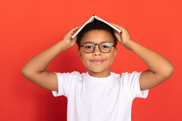 Portrait of happy schoolboy wearing glasses holding book on head. Mixed race preteen boy wearing...