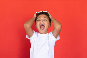 Portrait of frustrated preteen boy holding book and shouting. Mixed race child wearing white T-shirt looking at camera and screaming against red background. Stress concept