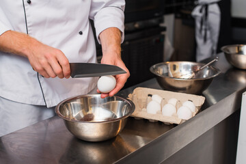 cropped view of chef in uniform holding knife near raw egg above bowl while cooking in kitchen.