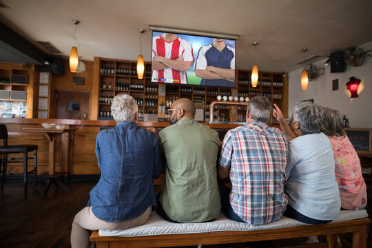 Diverse senior friends in bar watching tv with football match on screen