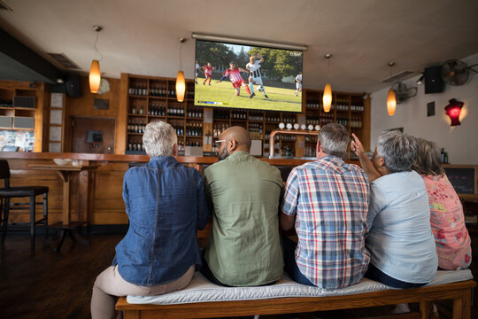 Diverse senior friends in bar watching tv with football match on screen