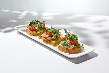 Aesthetic composition with shrimp bruschetta on white background with shadows from flowers. Italian bruschetta with shrimp, tomatoes and arugula on fine dining in summer. Elegant menu concept.