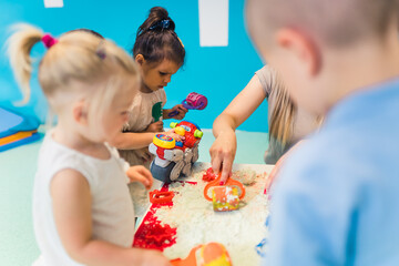 Relaxing sensory play with moldable kinetic sand at nursery school. Toddlers with their teacher having fun around the table using different tools for sculpting sand such as colorful and textured - obrazy, fototapety, plakaty