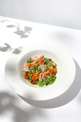 Elegant salad with baked salmon in sauce with vegetables on white table with harsh shadows. Salmon salad with radish on white background with shadows of leaves. Summer salad with roasted trout.